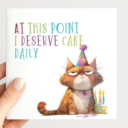 Funny Cat Birthday Card by Whimsi Lilley featuring a humorous cat illustration and the phrase "At this point I deserve cake daily."