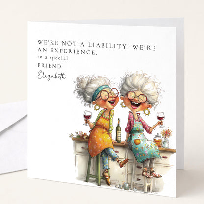 Funny personalised birthday card for friend with two older women laughing over red wine in kitchen captioned Not a Liability An Experience