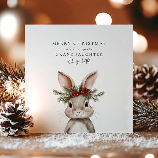 May include: A Christmas card featuring a watercolour illustration of a grey bunny wearing a wreath of greenery and red berries. The card reads "MERRY CHRISTMAS to a very special GRANDDAUGHTER Elizabeth". The card is on a wooden surface with pinecones.