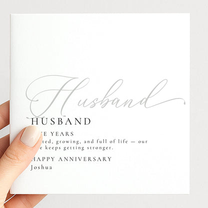 a hand holding a card with the word "Husband" written on it. The card has a definition of a husband and a happy anniversary message.