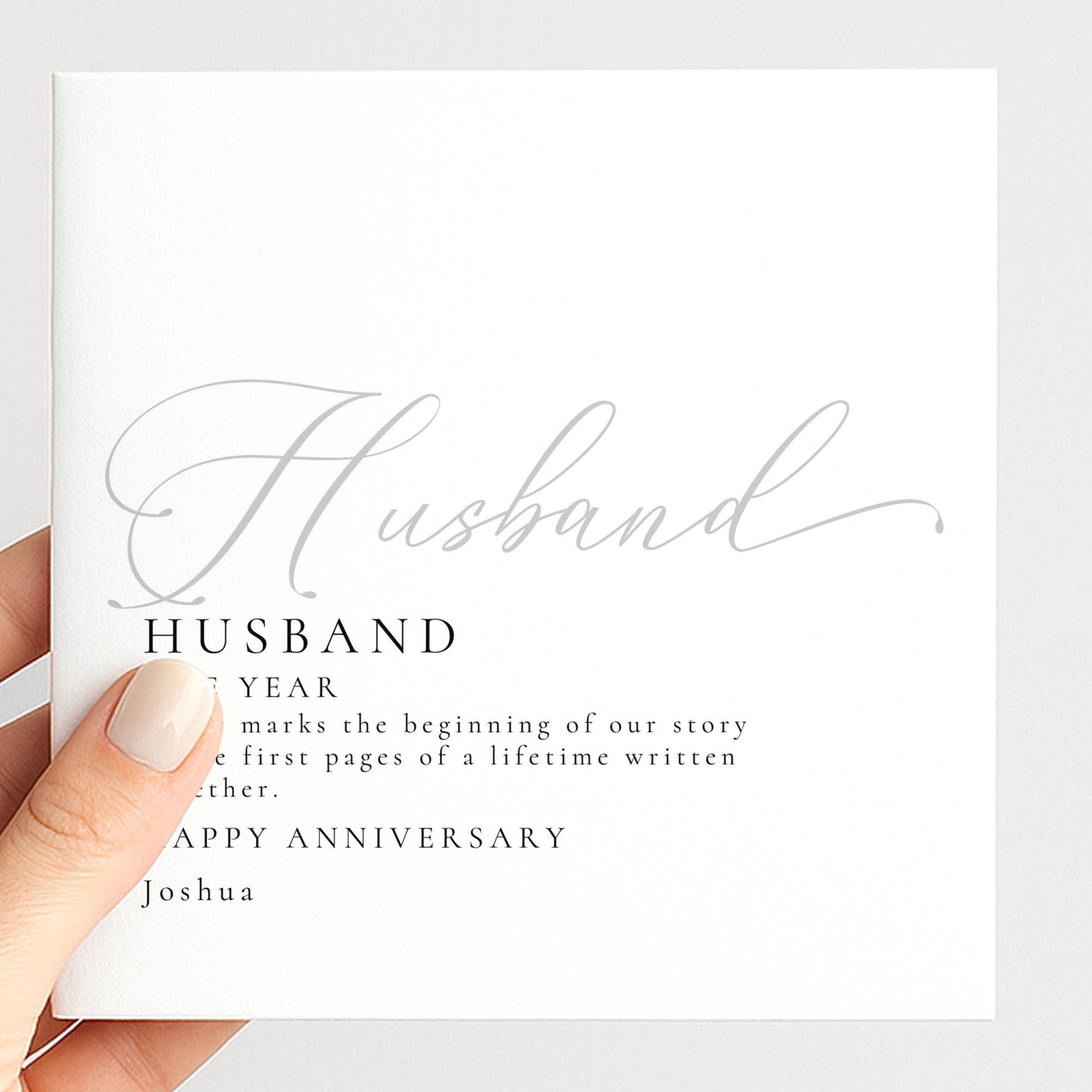 A hand holding a white card with the text "Husband" and "Happy Anniversary Joshua" printed on it.