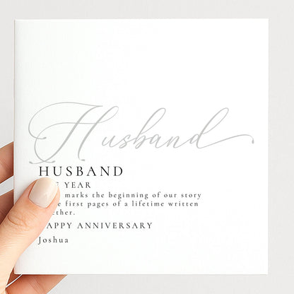 A hand holding a white card with the text "Husband" and "Happy Anniversary Joshua" printed on it.