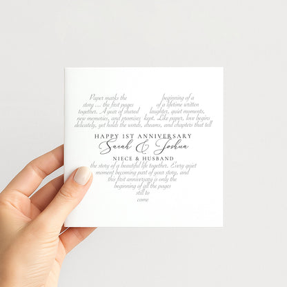 a hand holding a white card with text on it. The card appears to be a wedding invitation or anniversary card, with the text celebrating the first anniversary of a couple.